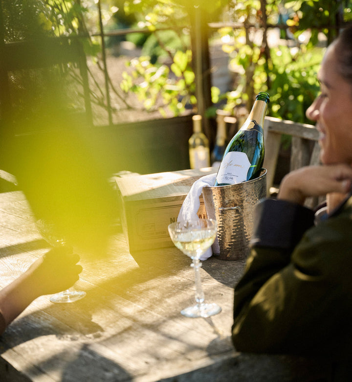 Meghan sitting at a table with a bottle of As ever Brut and a glass outdoors.