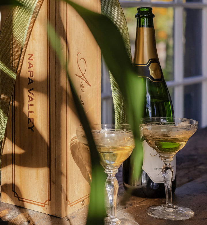 Bottle of As ever Brut, two glasses with champagne, and an As ever wooden crate with the text "Napa Valley" on a table